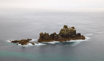 Long exposure rocks This landscape photograph captures a long exposure view of rugged rocks rising from the sea off the coast of Cornwall. The image shows the tranquil surface of the water, smoothed out by the long exposure technique, with the jagged cliffs and rocky outcrops contrasted against the gentle hues of the evening sky. Taken in early summer, the light suggests late afternoon approaching evening, illuminating the textures of the cliffs and the surrounding turquoise sea. The scene emphasizes the geological character of the Cornish coastline, with the isolated rocky formations standing prominently as the main subject in the composition.